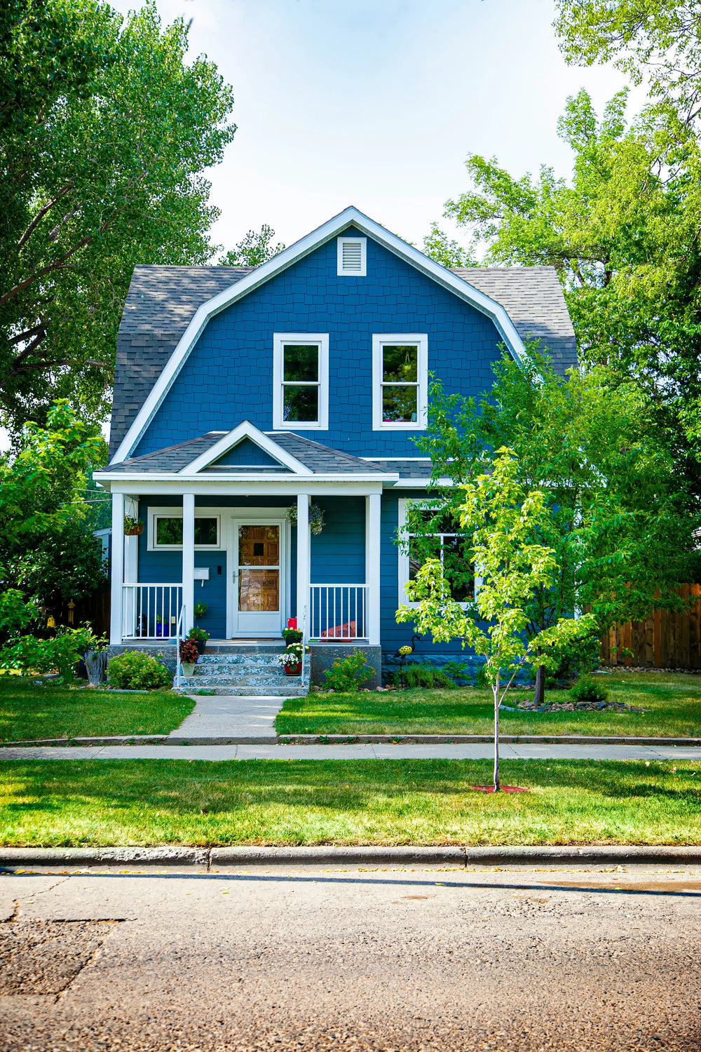 A welcoming two-story blue house with a gambrel roof, white front porch, and lush green yard, representing Mora House Plans